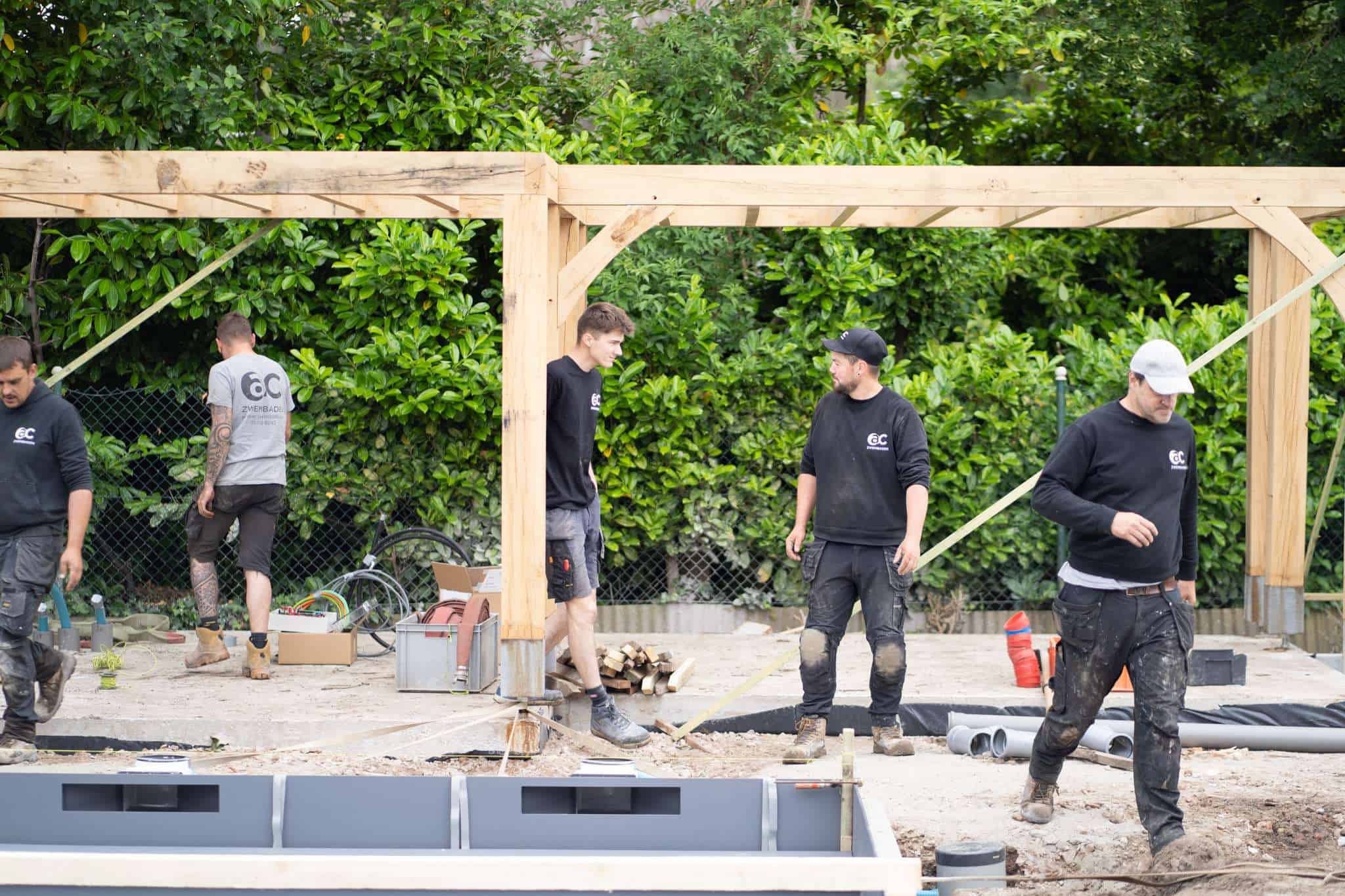 Medewerkers van AC Zwembaden in bedrijfskleding tijdens de constructie en installatie van een zwembad bij een houten bijgebouw of pergola.