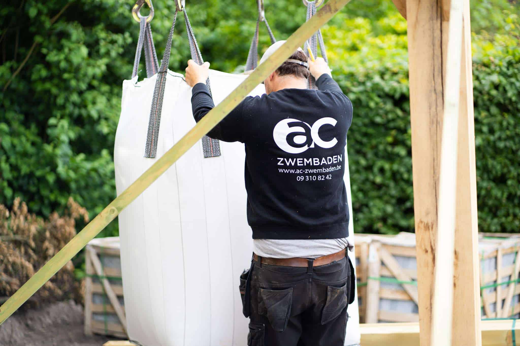 Medewerker van AC Zwembaden van achteren gezien bij het begeleiden en lossen van een grote big bag op de werf.